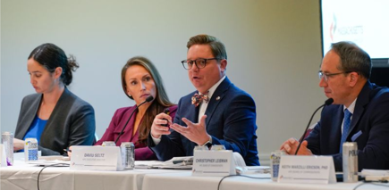 Photo of David Seltz speaking into a microphone giving the research presentation. Three other individuals are seated next to him listening to the presentation.
